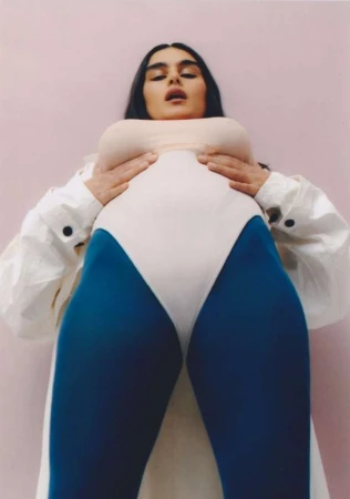 Low angle portrait of a woman wearing blue leggings, white bodysuit, and open white jacket against a pink backdrop by Zoe Natale Mannella