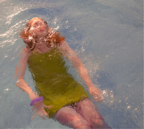 Woman in green dress floats in a pool with clear water; a purple bracelet adorns her wrist as she peacefully relaxes in the water.