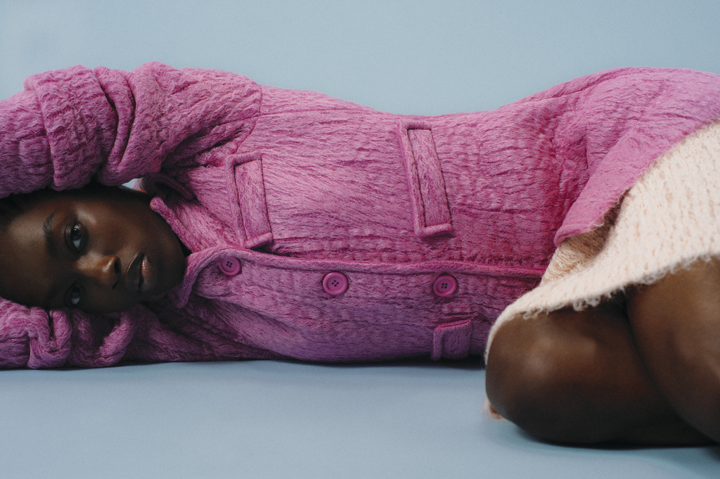 Woman with dark hair and brown eyes lies with hands behind her head, wearing pink buttoned jacket against light blue backdrop. 
