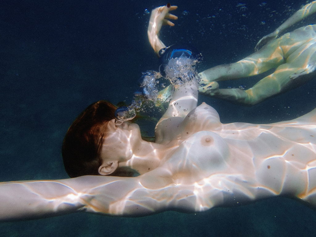 Two naked women swimming underwater, one with a diving mask, in dark blue water with light shining through the surface above them.