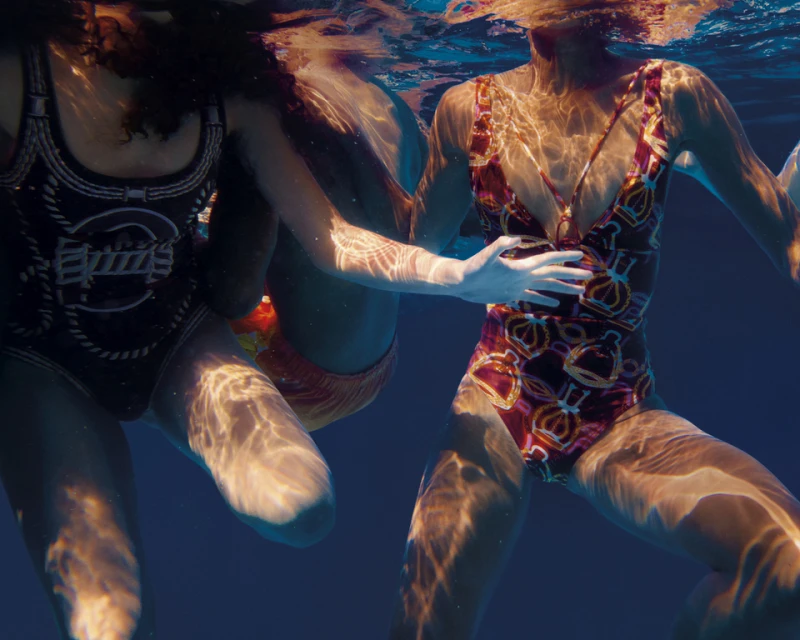 Two women in patterned swimsuits are partially submerged in blue water with light reflecting off the surface and illuminating their bodies.
