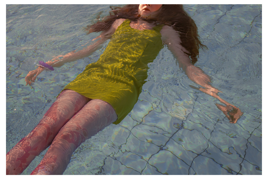 Woman in a yellow dress floats in a patterned pool, the water distorting her figure in a serene and sunlit scene.