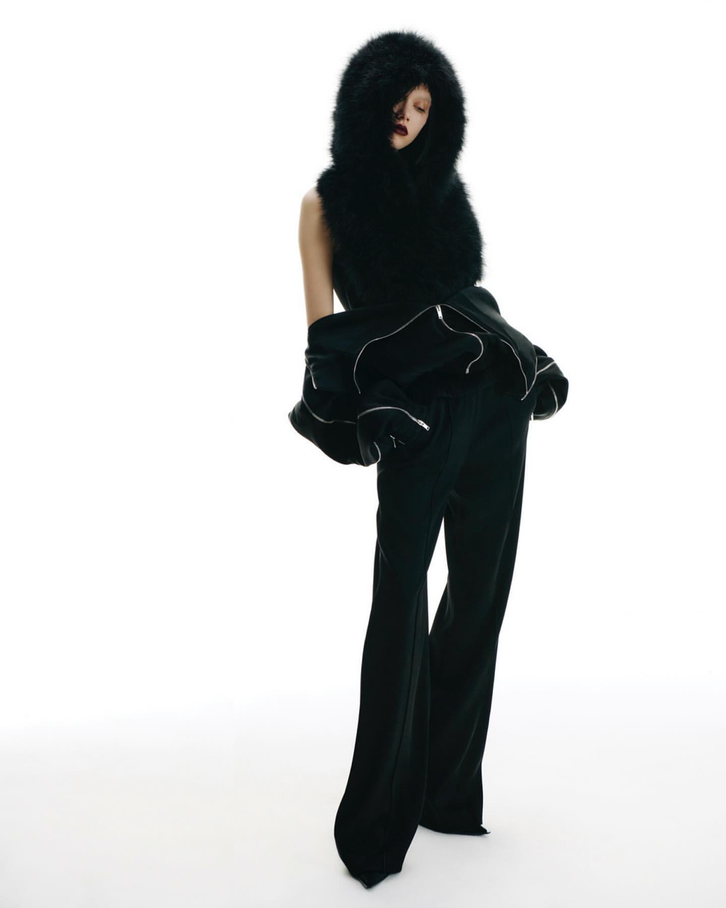 Woman in dark flared pants and jacket with a large faux fur hood. Studio shot against white backdrop. Fashion forward clothing design.
