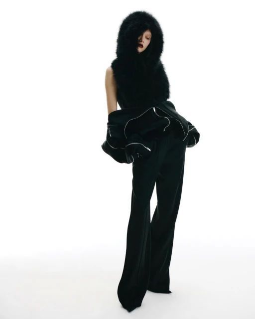 Woman in dark flared pants and jacket with a large faux fur hood. Studio shot against white backdrop. Fashion forward clothing design.