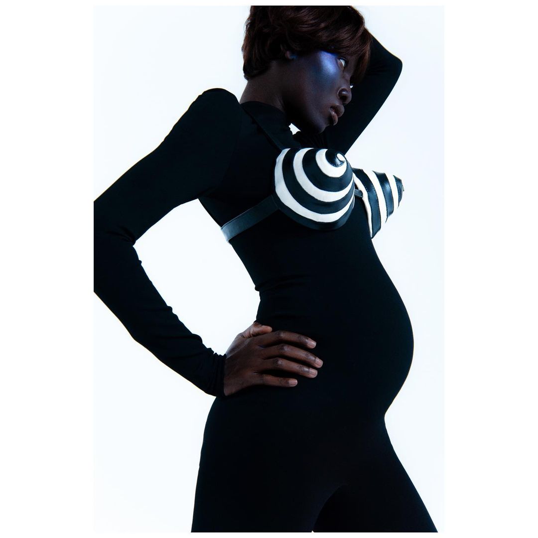 A pregnant African-descent woman poses in a black bodysuit with a spiral-patterned bra, showcasing maternity fashion against a white backdrop.