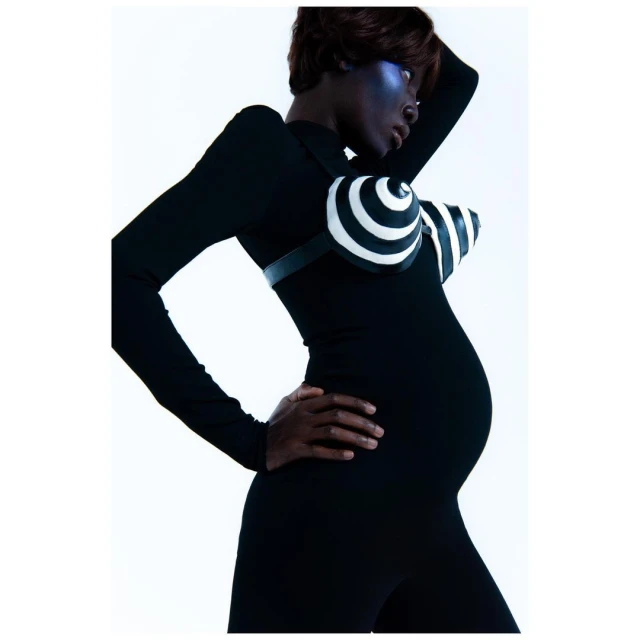 A pregnant African-descent woman poses in a black bodysuit with a spiral-patterned bra, showcasing maternity fashion against a white backdrop.