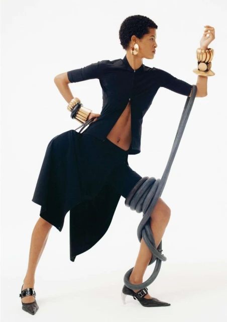 Fashion shot of a Black woman in a black shirt and skirt, posing dynamically with a gray rope, wearing gold earrings and bracelets.
