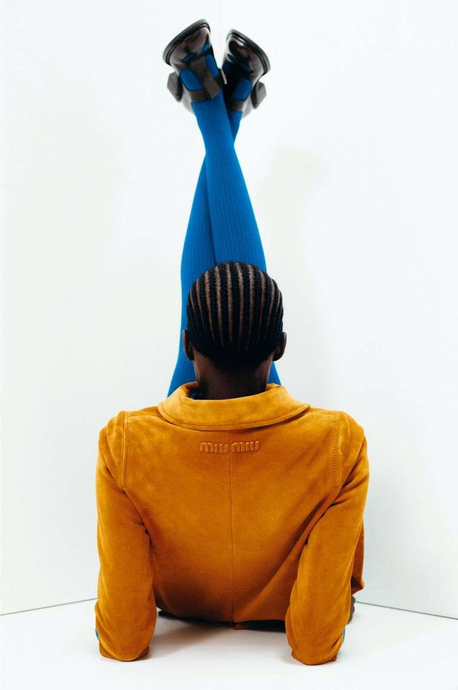 Woman poses with legs elevated wearing blue tights, black shoes, and tan jacket against a white corner in a fashion editorial.