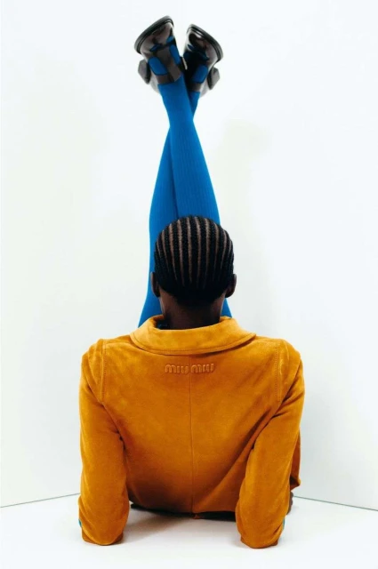 Woman poses with legs elevated wearing blue tights, black shoes, and tan jacket against a white corner in a fashion editorial.