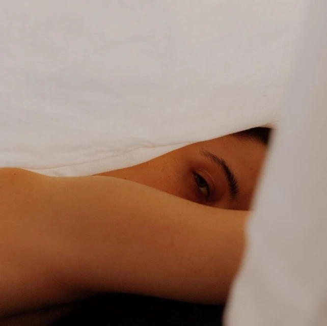 Close-up shot of an adult female lying under a white sheet, with her face partially visible and arm covering part of her face.