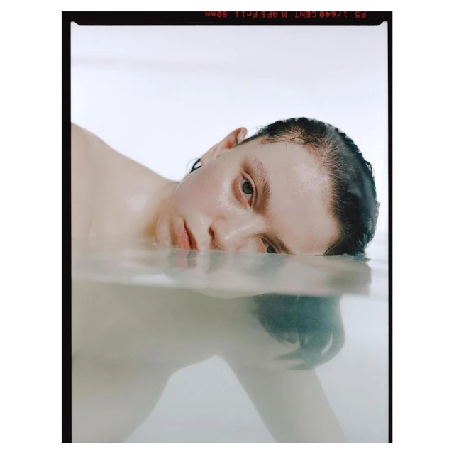 Portrait of a fair-skinned young adult with wet dark hair partially submerged in water, eye-level angle, and a clean white background.