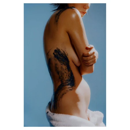 Studio shot of a person from the waist up with dark cosmetic clay on their skin, posing in front of a light blue background and wearing a white towel.