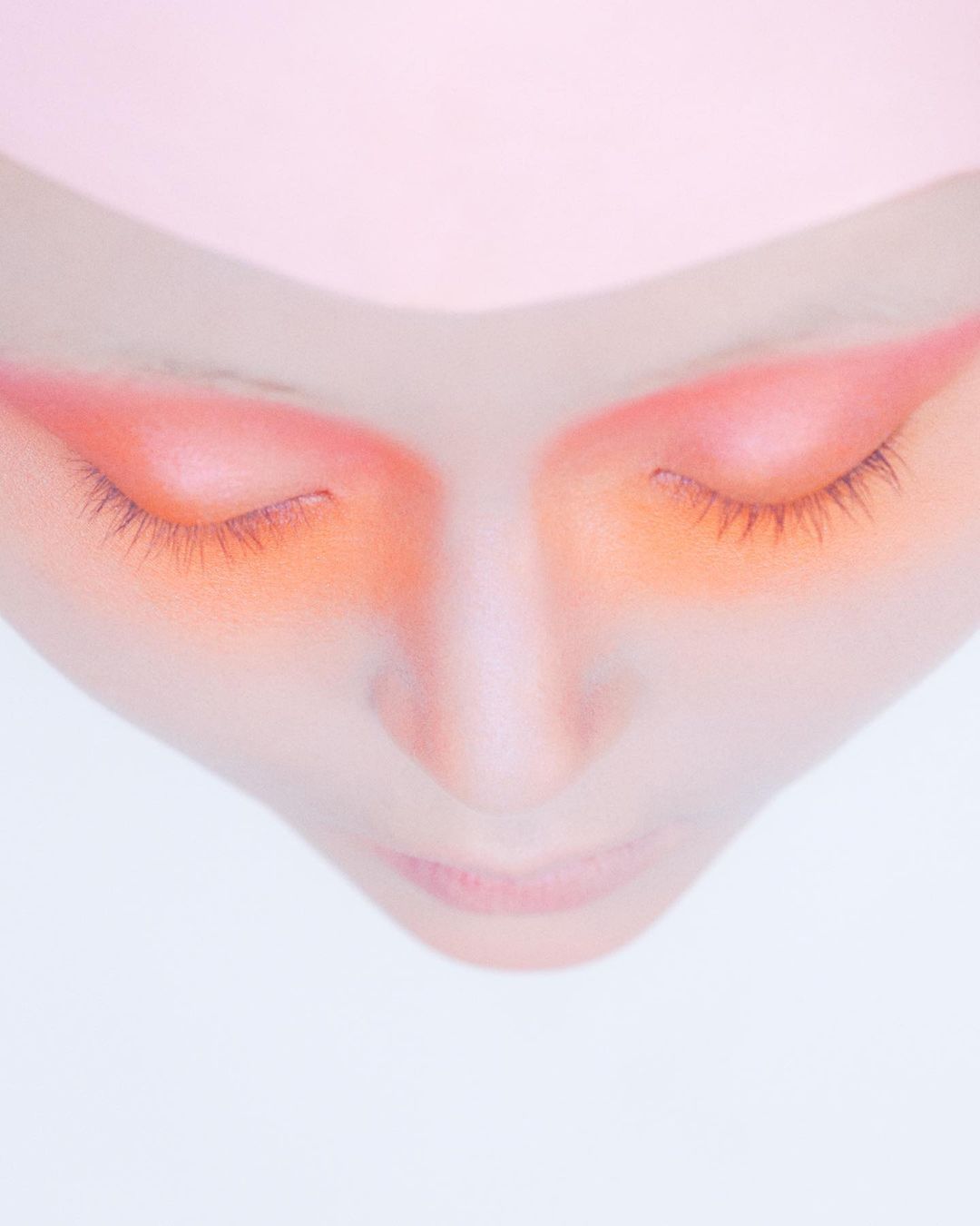Close-up of fair-skinned person with closed eyes and orange eye shadow against a light pink and white background in a portrait orientation.