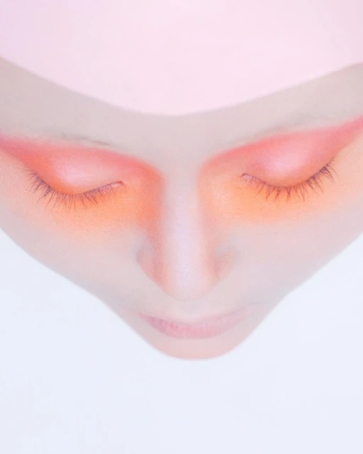 Close-up of fair-skinned person with closed eyes and orange eye shadow against a light pink and white background in a portrait orientation.