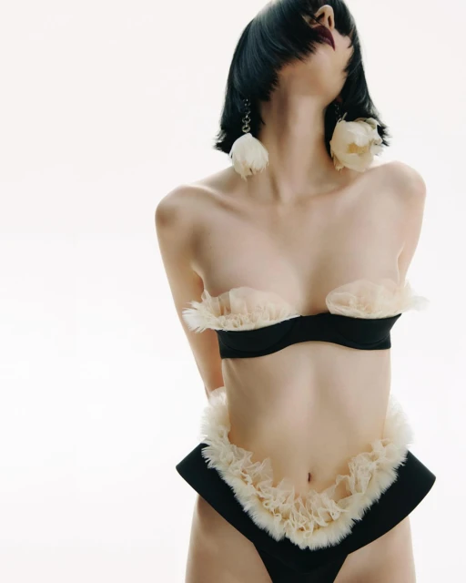 A female model poses in black lingerie with white ruffled trim against a clean white background, accentuating a minimalist aesthetic.