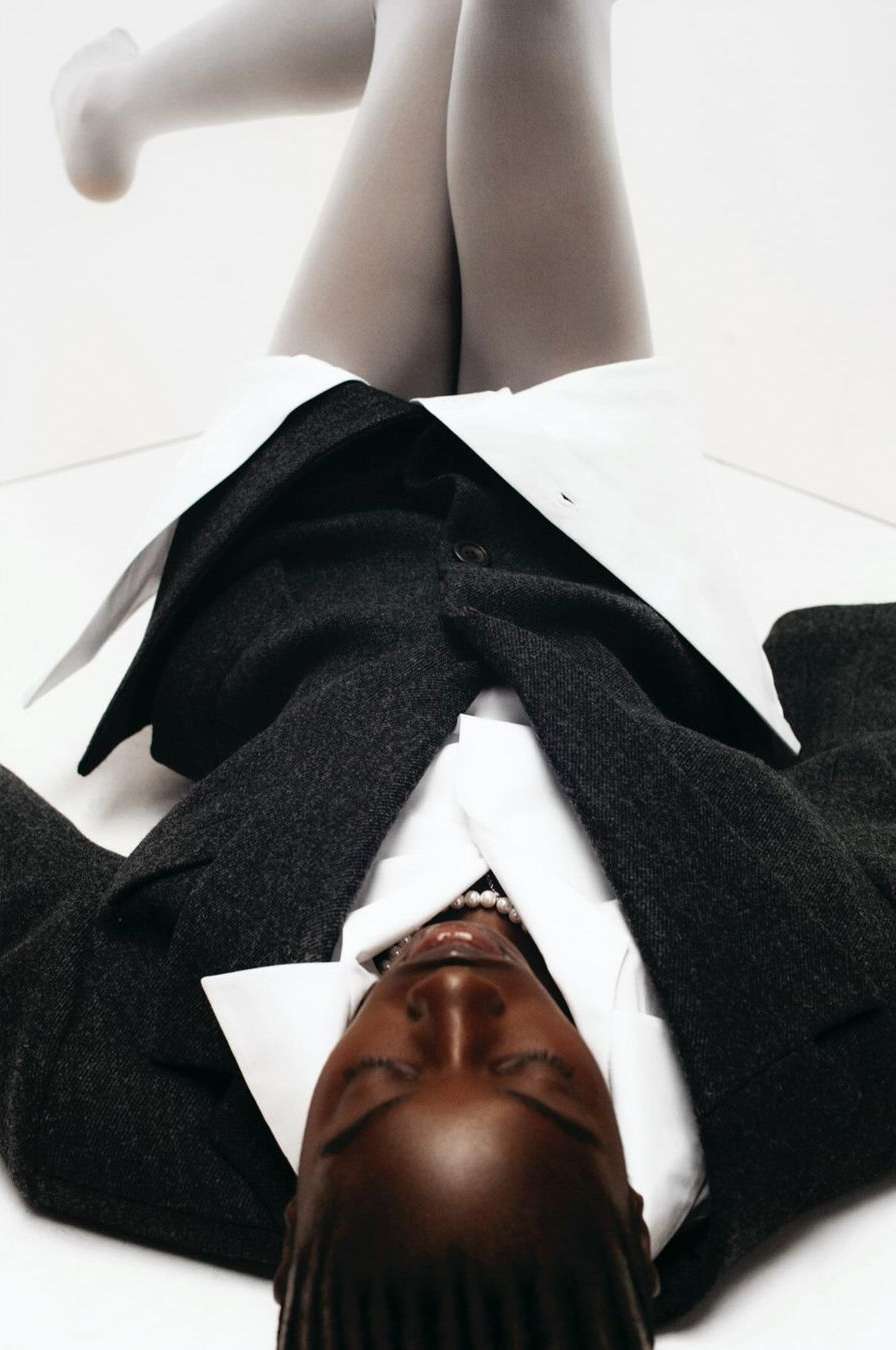 Fashion shot: woman lying on white backdrop wearing grey tights, a white collared shirt, pearl necklace and a gray jacket.