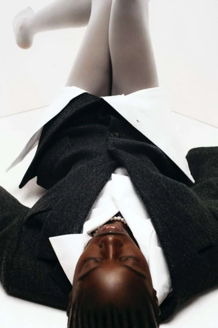Fashion shot: woman lying on white backdrop wearing grey tights, a white collared shirt, pearl necklace and a gray jacket.