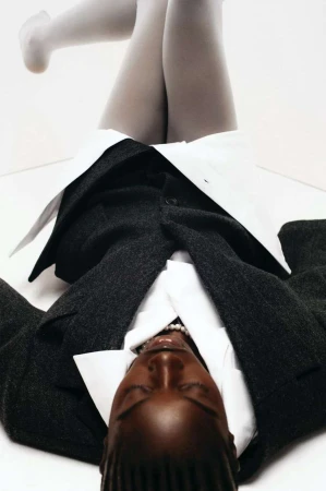 Fashion shot: woman lying on white backdrop wearing grey tights, a white collared shirt, pearl necklace and a gray jacket.