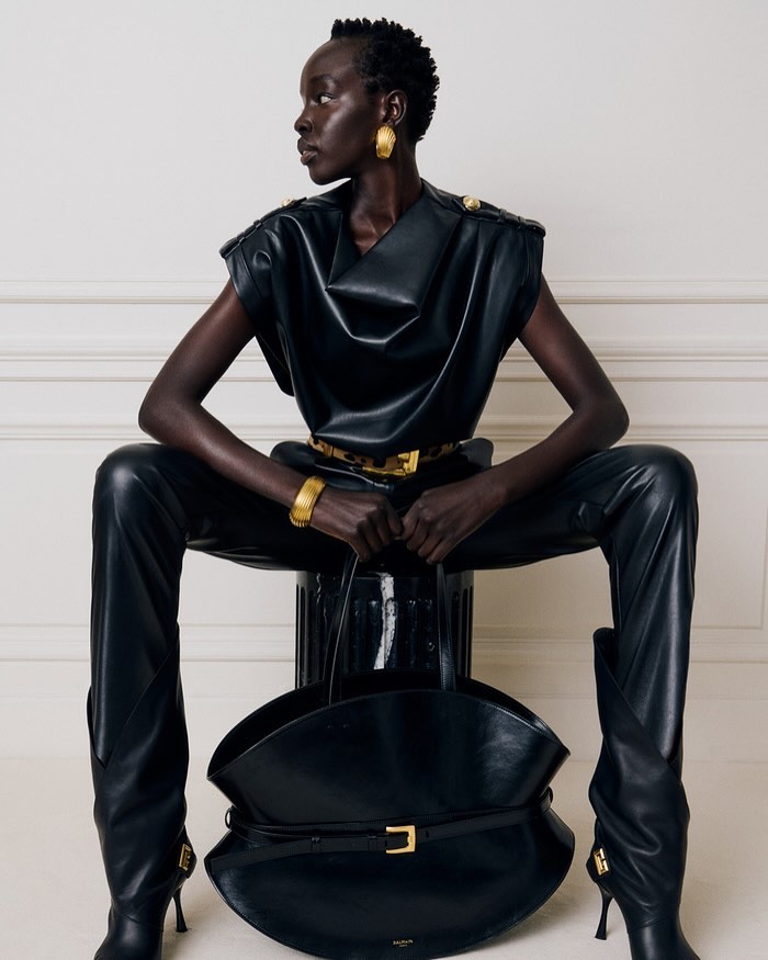 Fashion model in black leather top and pants sits with large black purse, accessorized with gold bracelet and earrings, neutral background.