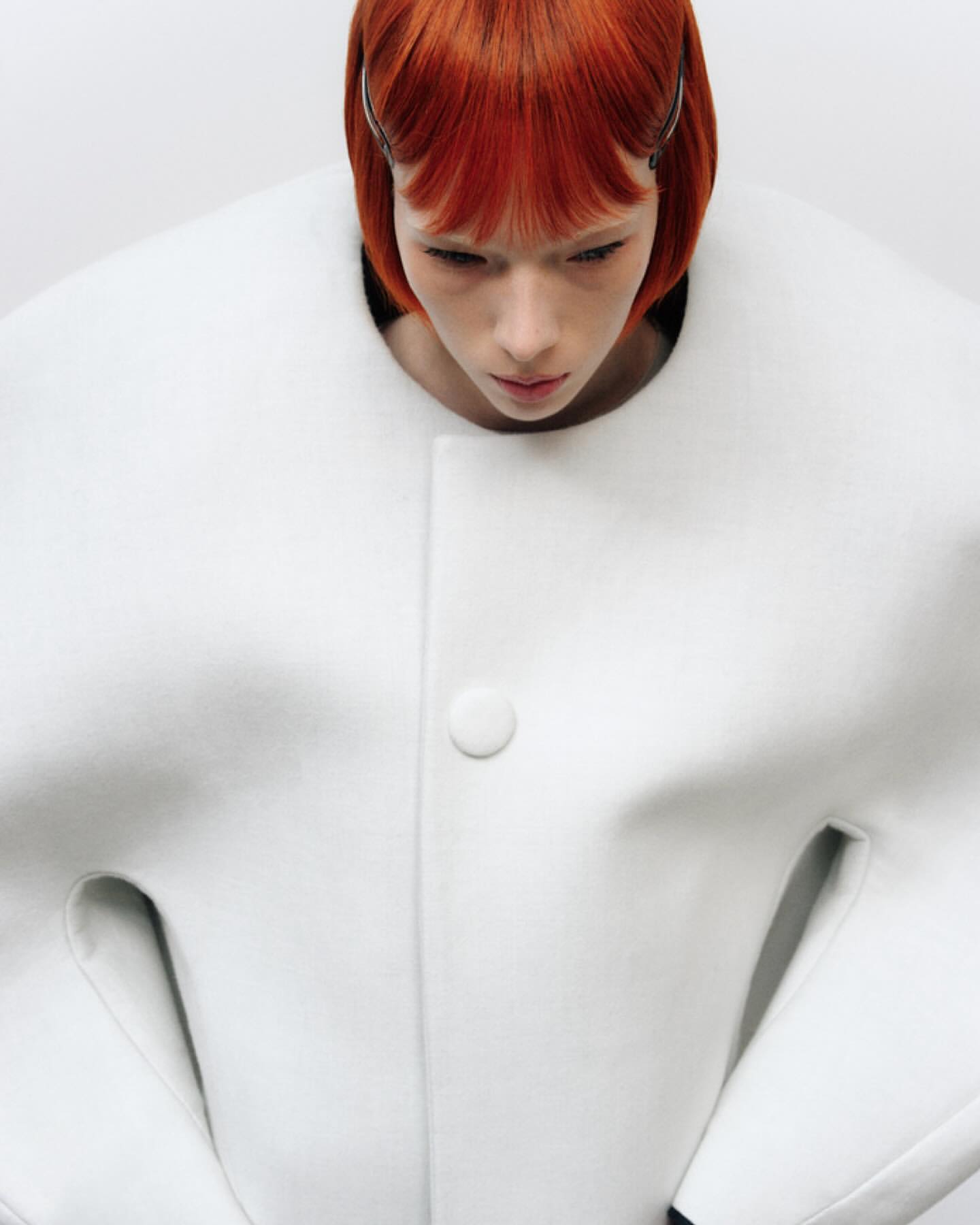 Fashion portrait: a pale-skinned woman with orange bob-cut hair wearing a white coat with rounded collar, shot against a white backdrop.