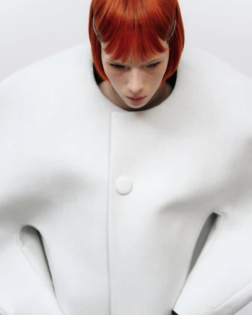 Fashion portrait: a pale-skinned woman with orange bob-cut hair wearing a white coat with rounded collar, shot against a white backdrop.