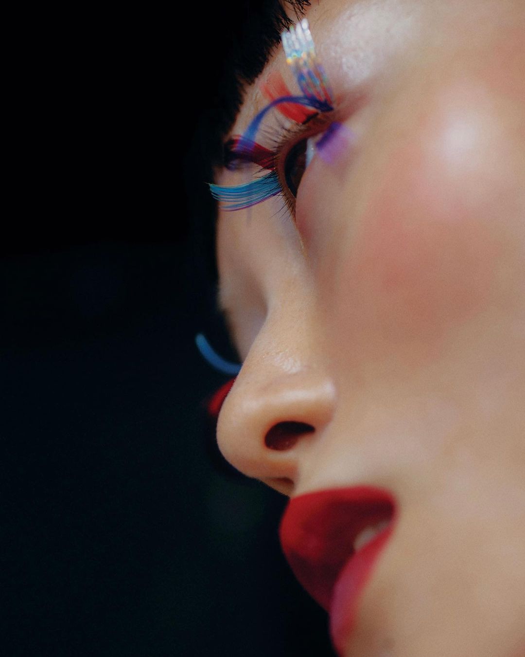 Close-up of a person's face with colorful false eyelashes, red lipstick, and fair skin against a stark black background, beauty theme.