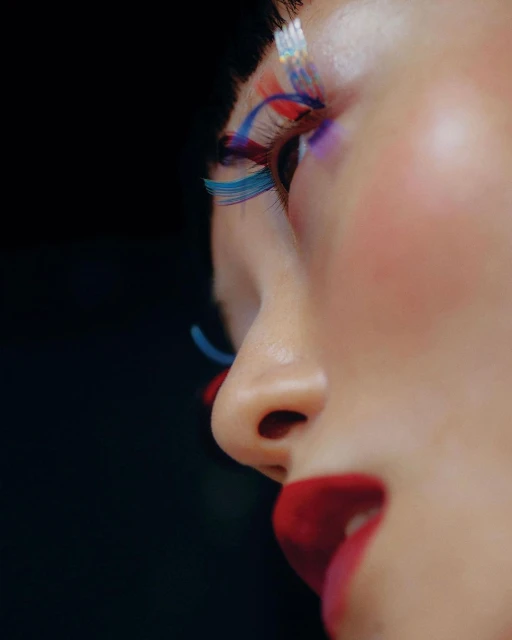 Close-up of a person's face with colorful false eyelashes, red lipstick, and fair skin against a stark black background, beauty theme.