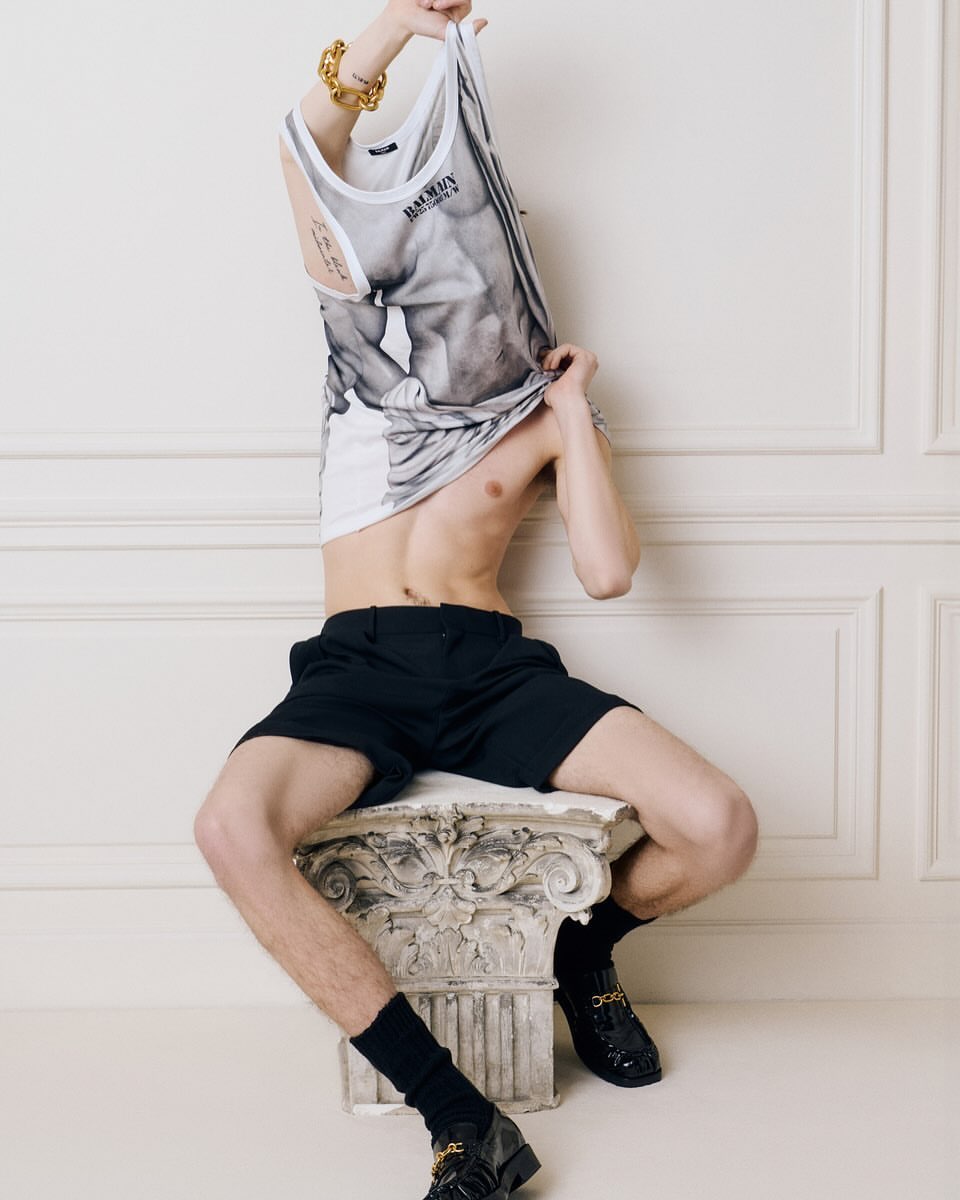 An adult male poses indoors, seated on a faux-Corinthian column, wearing black shorts and patent shoes, and pulling up a graphic-print tank top.