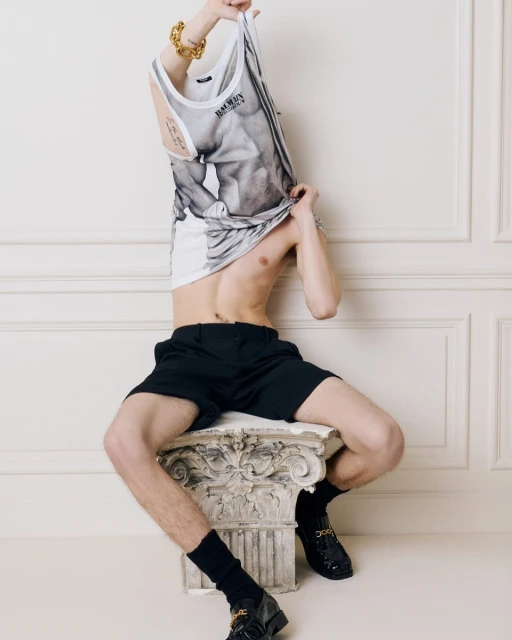 An adult male poses indoors, seated on a faux-Corinthian column, wearing black shorts and patent shoes, and pulling up a graphic-print tank top.
