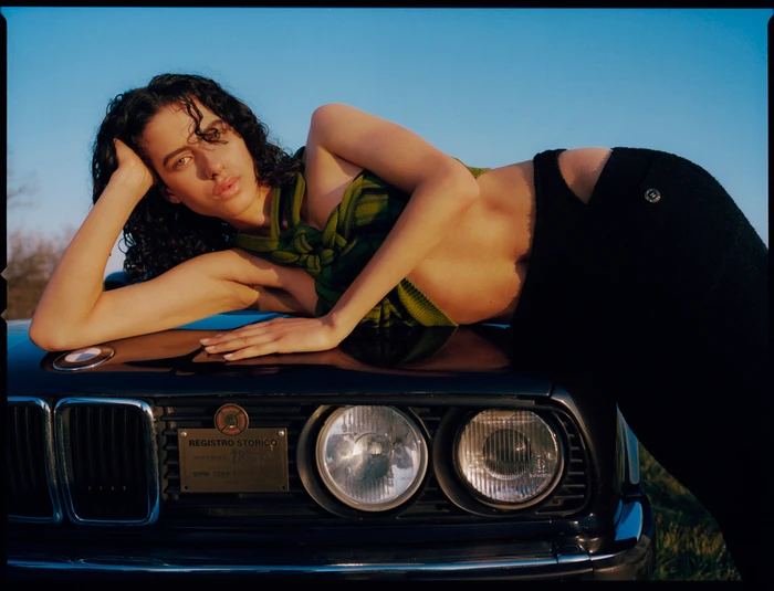 A woman reclining on the hood of a classic dark car against a clear blue sky, wearing a green top and dark pants.