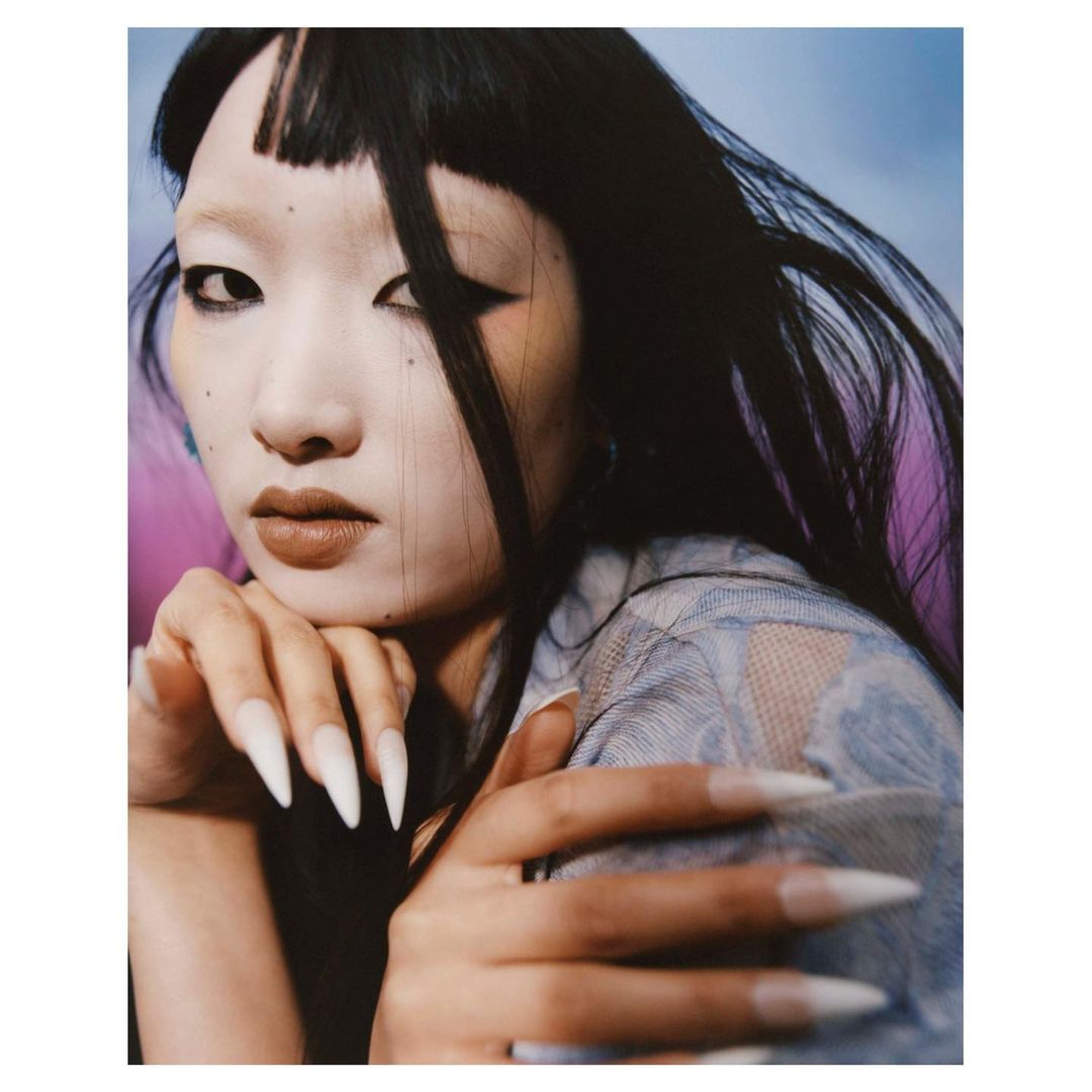 Close-up portrait of an East Asian woman with long dark hair, wearing a sheer blue top and featuring long, white-tipped fingernails.