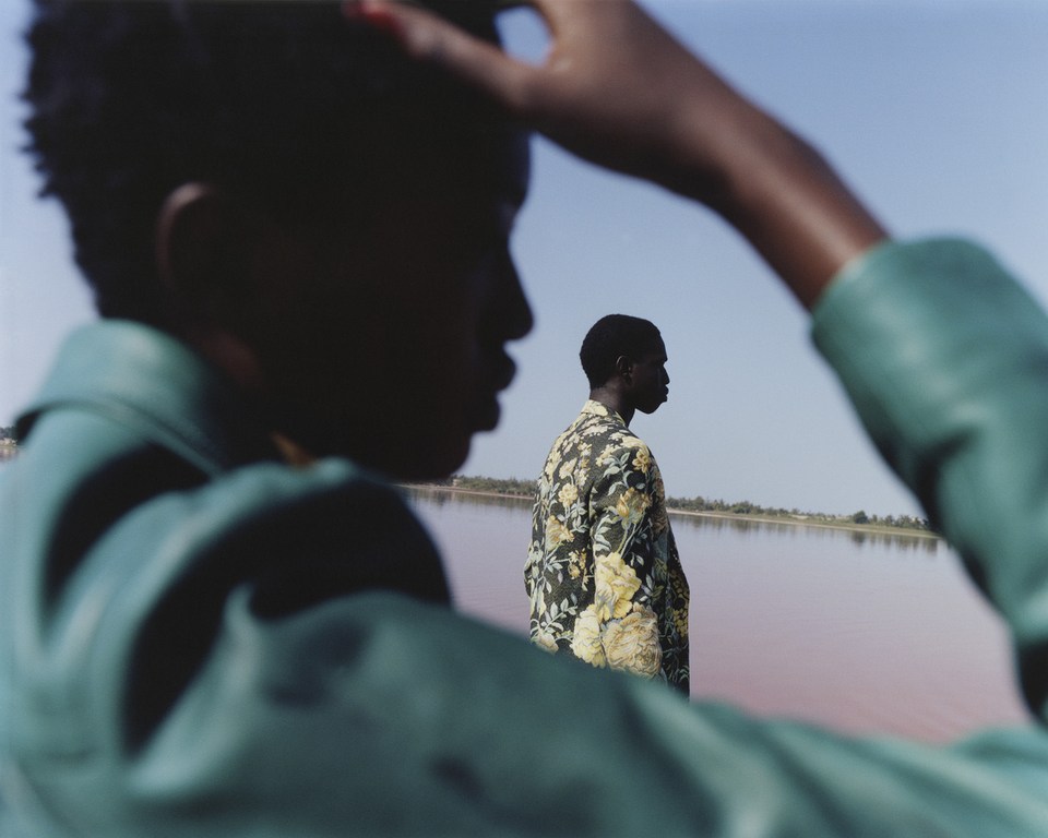 Two black men in patterned shirt and green jacket stand by the pink water of Lake Retba, Senegal, under a clear blue sky on a sunny day.