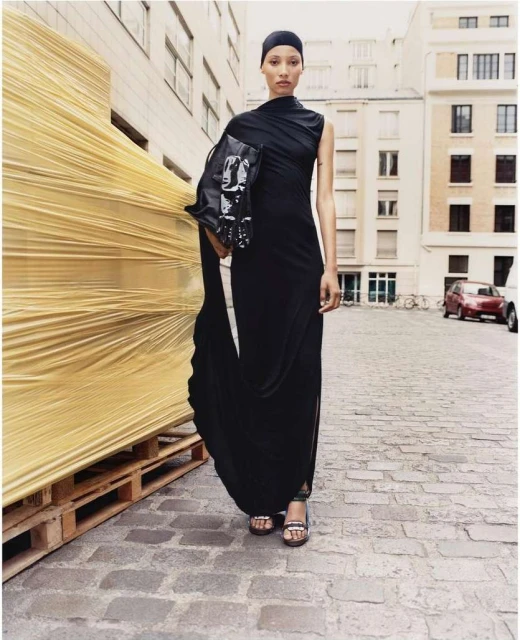 Elegant Black Woman in Designer Dress Posing on Cobblestone Street. Holding Clutch Bag. Building and Pallet Background.