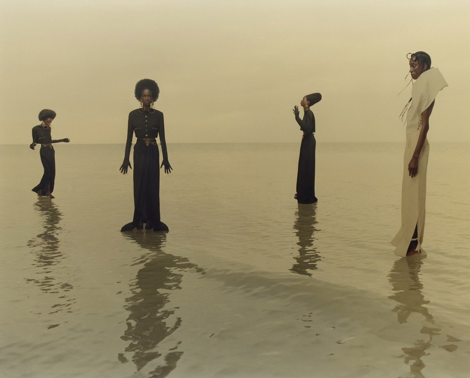Four women in long dresses stand in shallow water. The backdrop is a light-yellow sky. The figures cast reflections in the water.