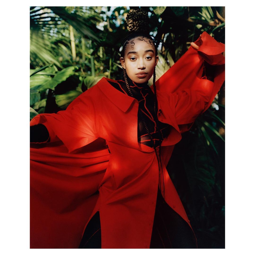 Young woman in red coat surrounded by lush green foliage. Portrait of a fashionable woman in a vibrant outdoor setting.