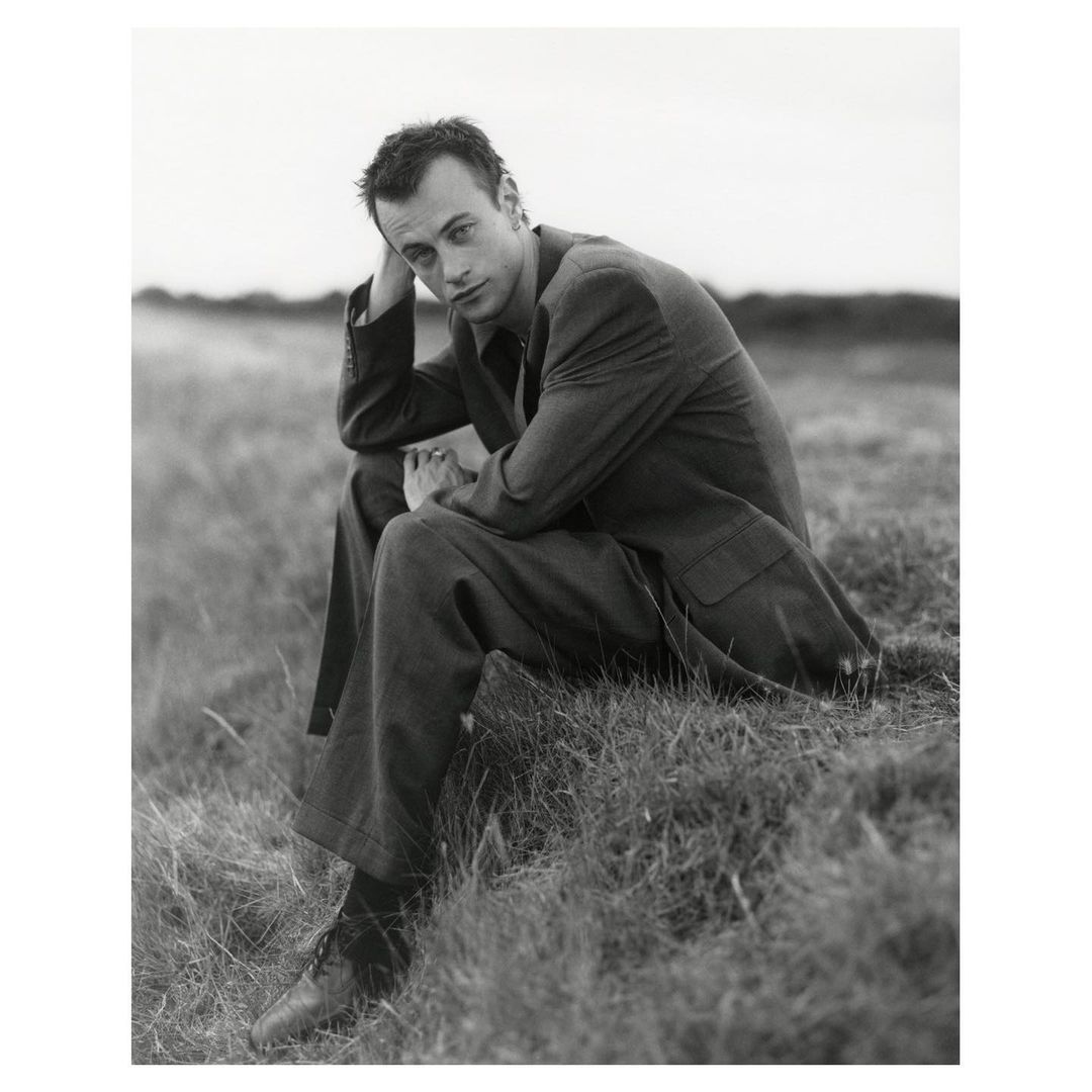 Black and white portrait of a man in a suit sitting in a grassy field looking at the camera with a neutral expression.
