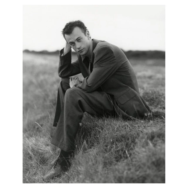 Black and white portrait of a man in a suit sitting in a grassy field looking at the camera with a neutral expression.