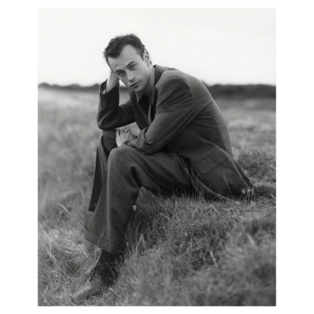 Black and white portrait of a man in a suit sitting in a grassy field looking at the camera with a neutral expression.