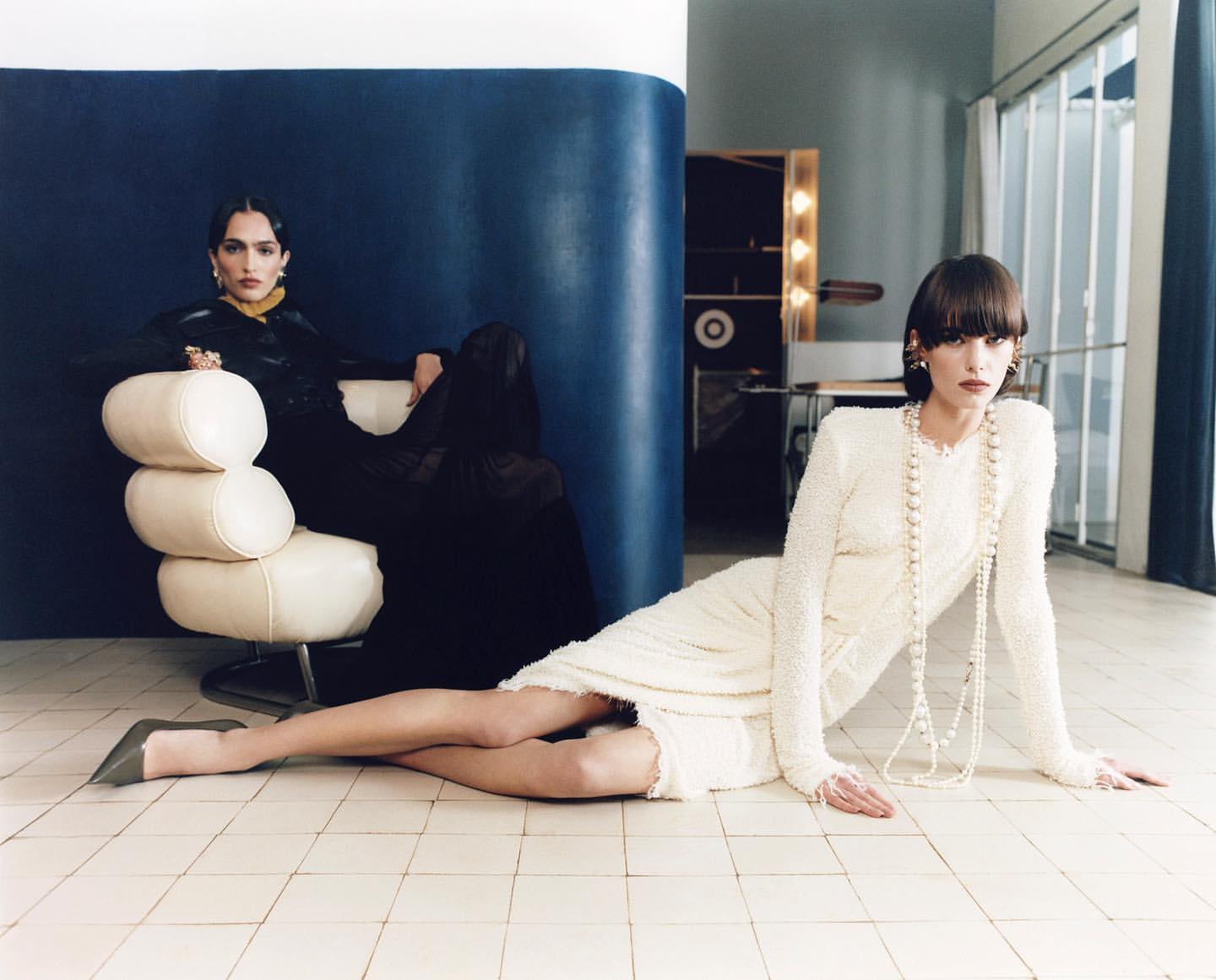 Two women are posing in a vintage-style indoor fashion shot with tiled flooring. One is sitting on a chair, the other laying down.