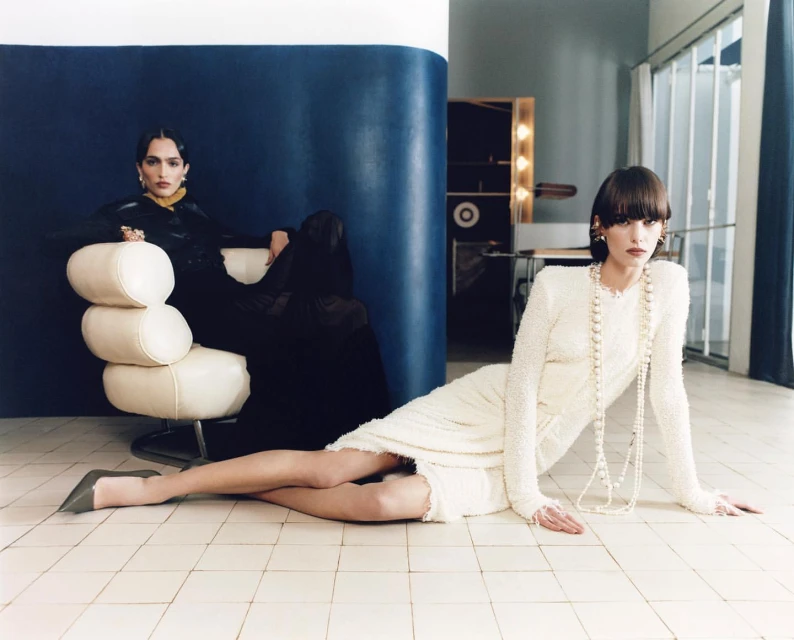 Two women are posing in a vintage-style indoor fashion shot with tiled flooring. One is sitting on a chair, the other laying down.