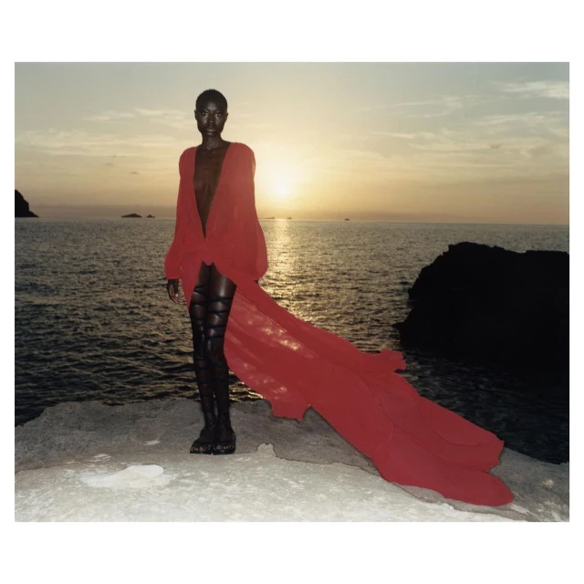 Woman in red robe poses on rocks near the ocean at sunset. Fashion editorial image with dark tones and a dramatic sky. 