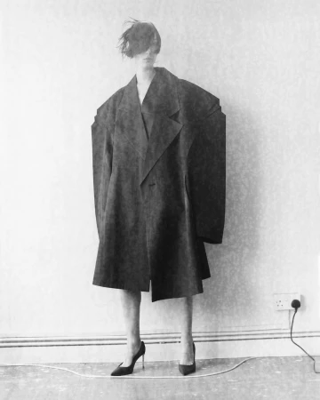 A person in a large black coat and black high heels poses against a wall with a power outlet in a stark, black and white fashion photo.