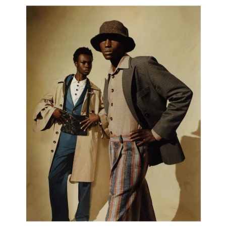 Two male models posing against a textured pale wall, wearing tailored patterned trousers, blazers, and bucket hats.