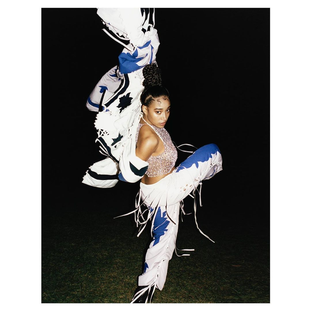 Woman in a shimmering top and decorated pants strikes a dynamic pose on a dimly lit lawn with a decorative white jacket overhead.