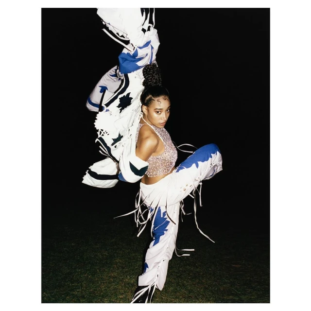 Woman in a shimmering top and decorated pants strikes a dynamic pose on a dimly lit lawn with a decorative white jacket overhead.