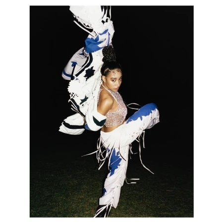 Woman in a shimmering top and decorated pants strikes a dynamic pose on a dimly lit lawn with a decorative white jacket overhead.