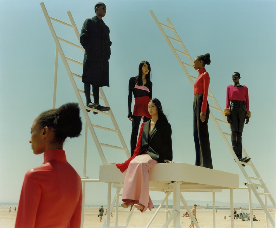 Fashion editorial on a beach featuring six adult models in stylish clothing posing on and around white ladders under a bright blue sky.