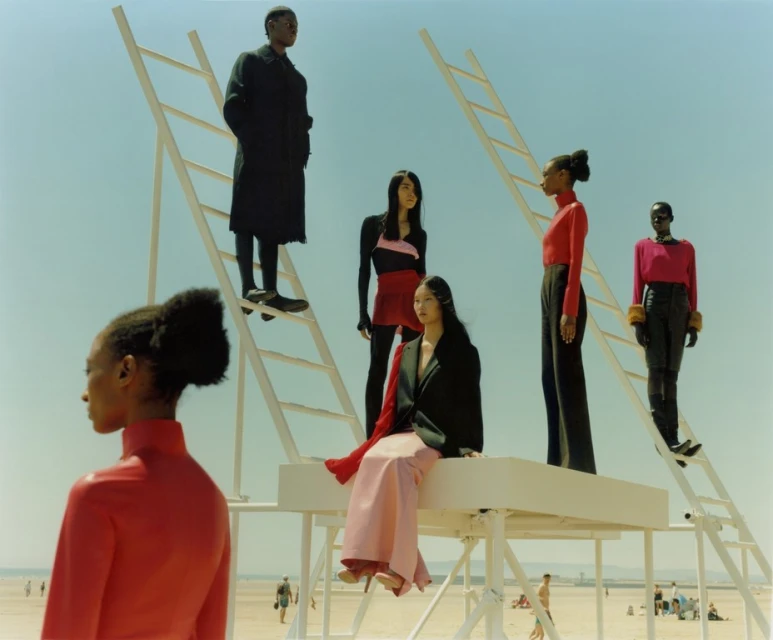 Fashion editorial on a beach featuring six adult models in stylish clothing posing on and around white ladders under a bright blue sky.
