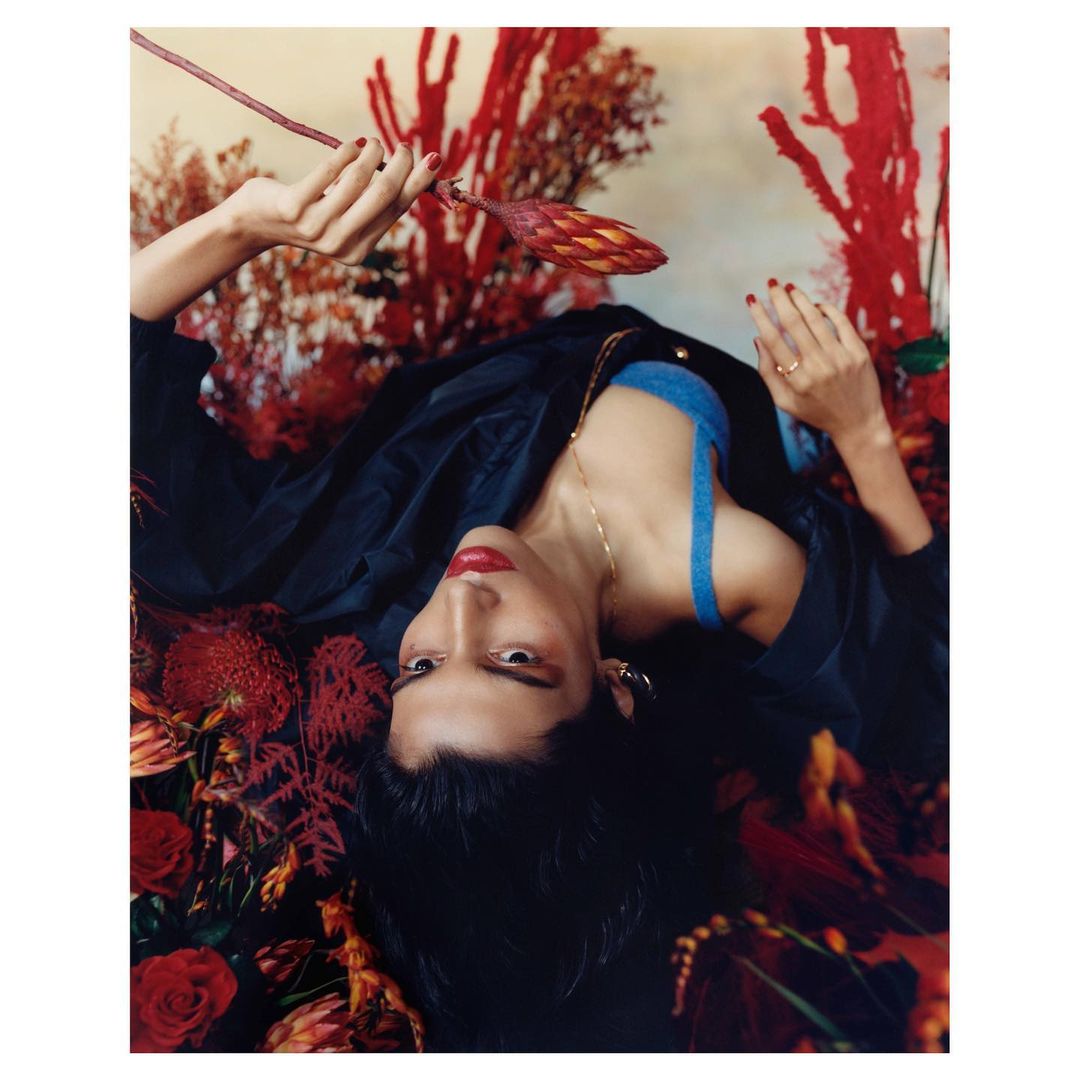 Portrait of a young woman with dark hair and red lipstick lying amongst red flowers, wearing a blue top and black jacket indoors.