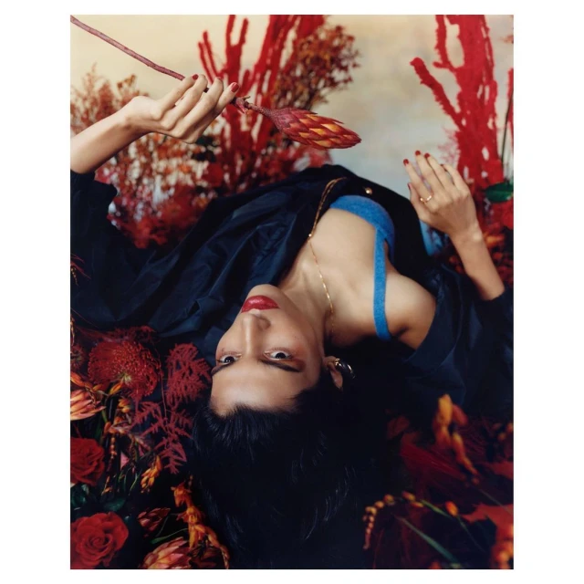 Portrait of a young woman with dark hair and red lipstick lying amongst red flowers, wearing a blue top and black jacket indoors.